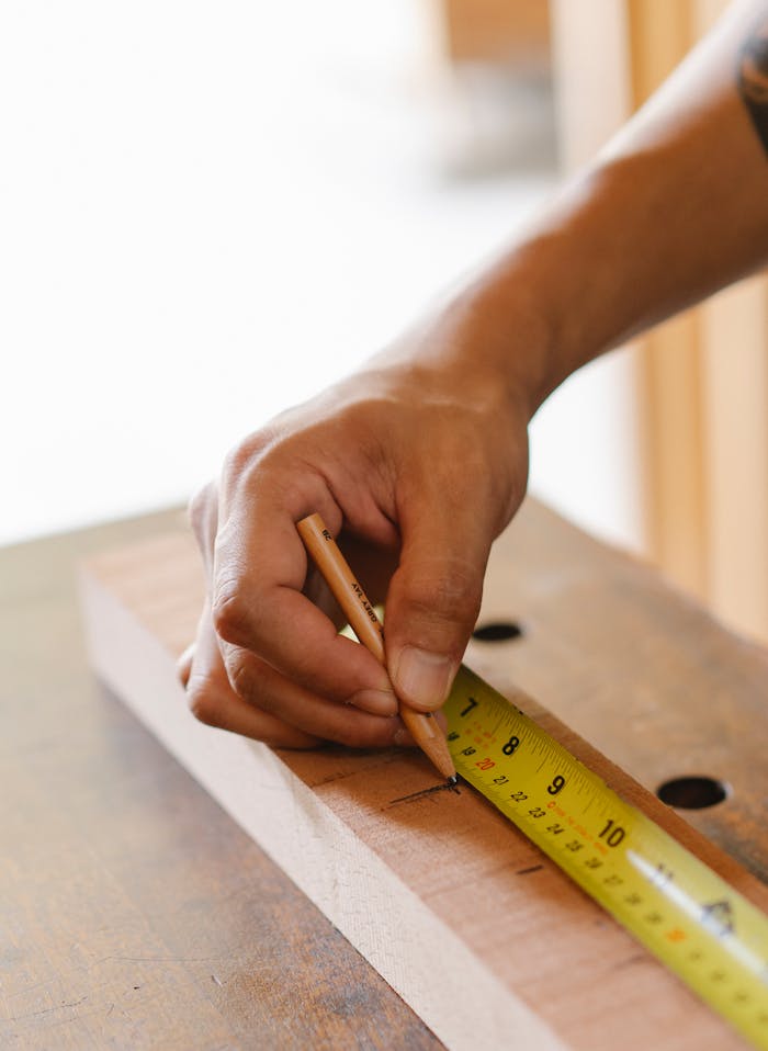 From above crop anonymous male joiner making sign with pencil measuring wooden plank while making handicraft in workshop